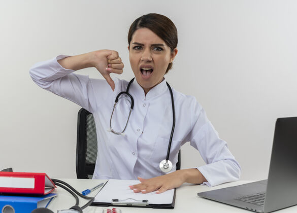 医疗悲伤的年轻女医生穿着医用长袍 手持听诊器坐在办公桌旁 拿着医疗工具在电脑上工作 拇指朝下 留着复印空间听诊器向下工具