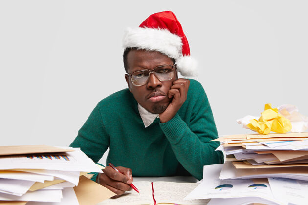 办公桌伤心不满意的男人捂着嘴唇 手贴着脸颊 戴着圣诞老人的帽子 在庆祝寒假前努力工作纸张凝视写作