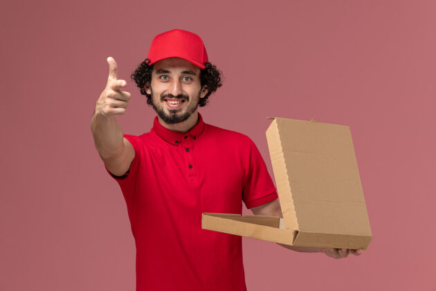 粉色正面图身穿红色衬衫和斗篷的男快递员拿着空的快递食品盒 微笑着站在粉红色的服务台上快递公司的员工服务观点信使