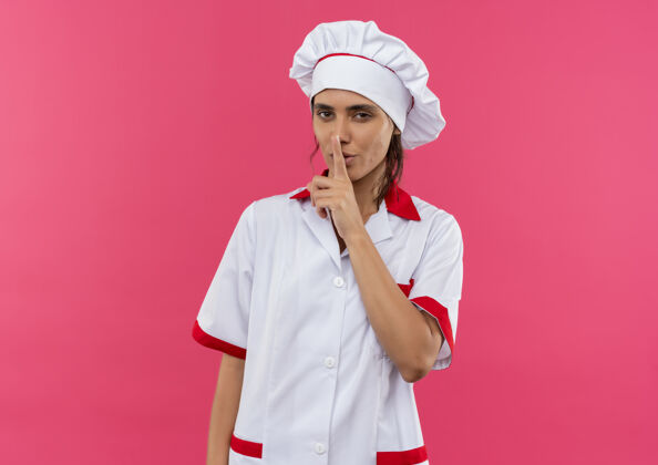年轻穿着厨师制服的年轻女厨师在粉色的墙壁上做了一个安静的手势厨师穿手势