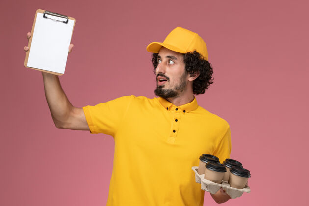 视图正面图：身穿黄色制服的男信使手拿棕色的咖啡杯 粉色墙上放着记事本记事本棕色工作