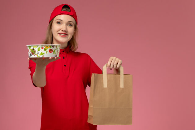 视图正面图：身穿红色制服 披风的年轻女快递员手持快递食品包和碗 微笑着站在粉红色的墙上快乐快递微笑