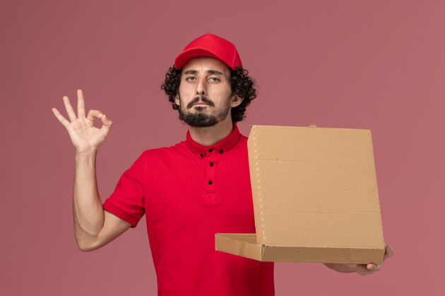 食物正面图：身穿红色衬衫和斗篷的男快递员拿着快递食品盒 在浅粉色的服务台上服务快递公司的员工持有男查看