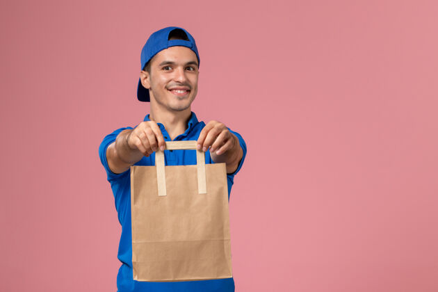 工作正面图：身穿蓝色制服和披风的年轻男性快递员 手上拿着一个纸包裹 放在粉红色的墙上男人手粉色