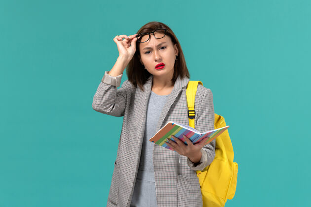 学校身穿灰色夹克 背着黄色背包 拿着抄写本的女学生在浅蓝色墙上的正视图女学生夹克课