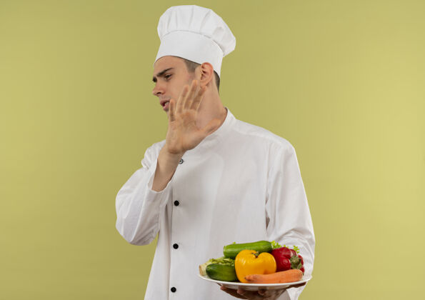 手势看着旁边那个穿着厨师制服的神经兮兮的年轻男厨师 手里拿着盘子里的蔬菜 在隔离的绿色墙上做着停车的手势制服厨师穿着
