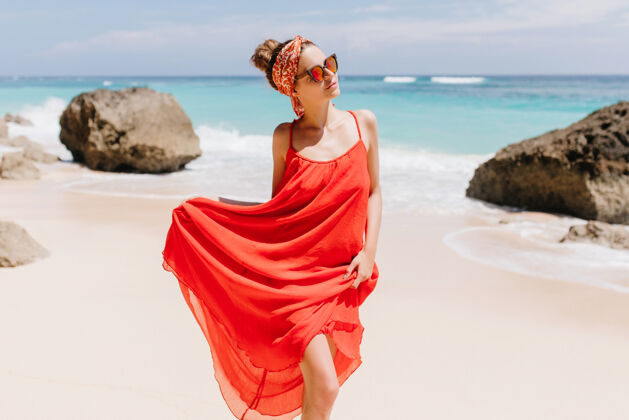 活动穿着红色长裙的可爱的年轻女子在有岩石的荒野海滩上摆姿势美丽的女孩在海边散步的户外镜头欢乐女人时尚